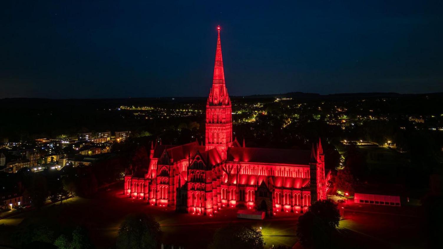 A drone shot of Salisbury cathedral at night light up completely red. Other city lights can be seen around it in the distance.