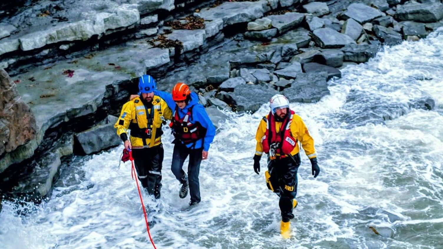 HM Coastguard and RNLI crew escort a person to safety using a rope as the waves crash around them.