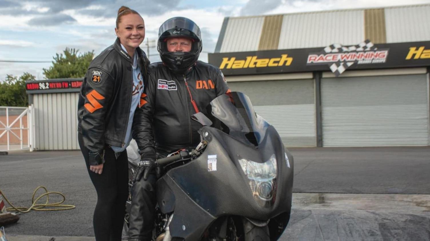 Danny's daughter stands next to him while he sits on his racing bike. He has the helmet on with visor up and wears a black jacket with the word DAN in orange. She wears a Santa Pod leather jacket too with the number 21 on a badge, over a blue t-shirt and black leggings.