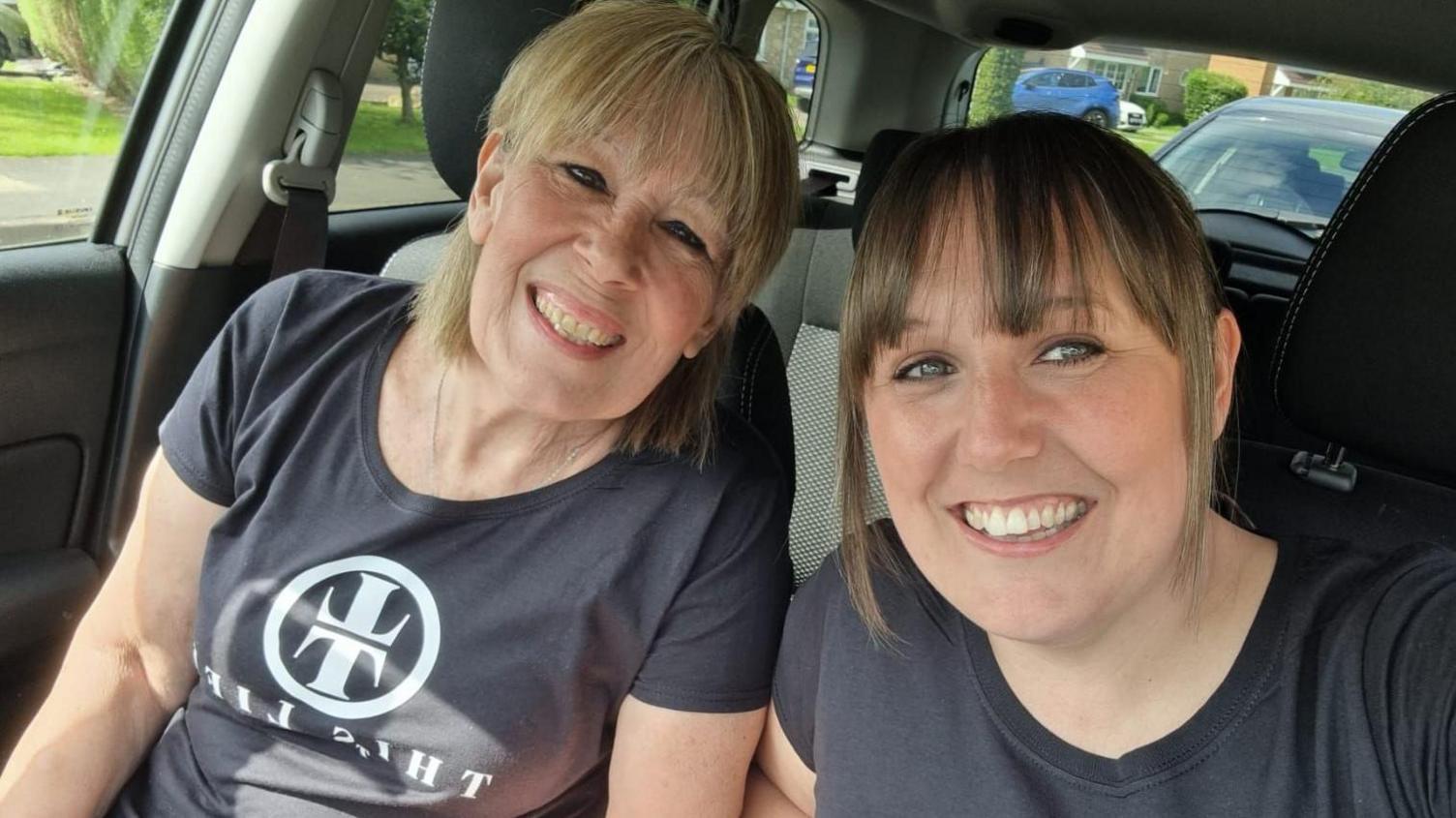 Lynda Sawyer and Becky Clough take a selfie while sitting in the front seats of a parked car. They are smiling into the camera and wearing black t-shirts with white Taylor Swift logos