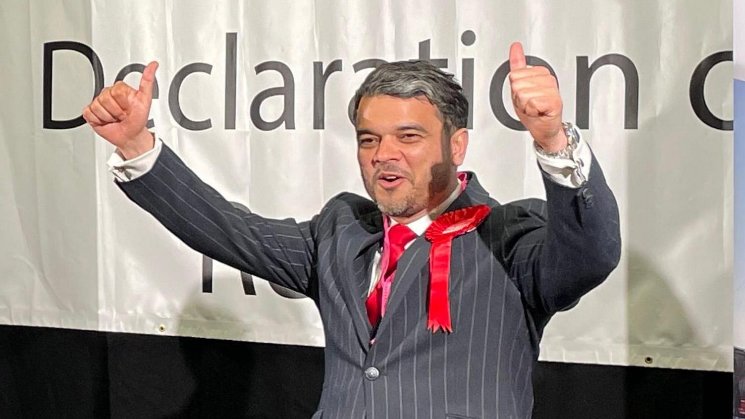 Cllr Iqbal wearing a pin stripe suit, red tie and Labour party rosette. Holding his hands in the air with thumbs up.  