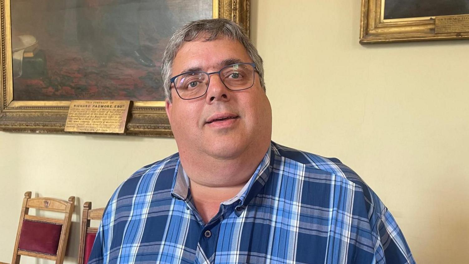 Councillor Richard Udall, a man wearing glasses and a shirt with blue checkered pattern on it. He is standing inside a cream-coloured room with gold-framed oil paintings hanging on the wall behind him.