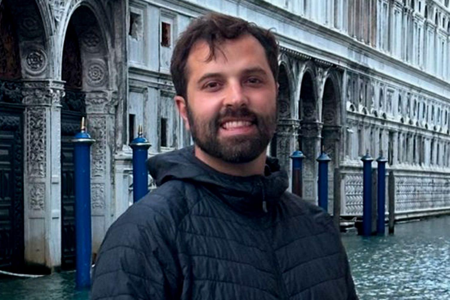 Justin Hodges has dark hair, a moustache and full beard. He is wearing a dark blue, padded, waterproof coat. He appears to be in Venice, by or on a canal.