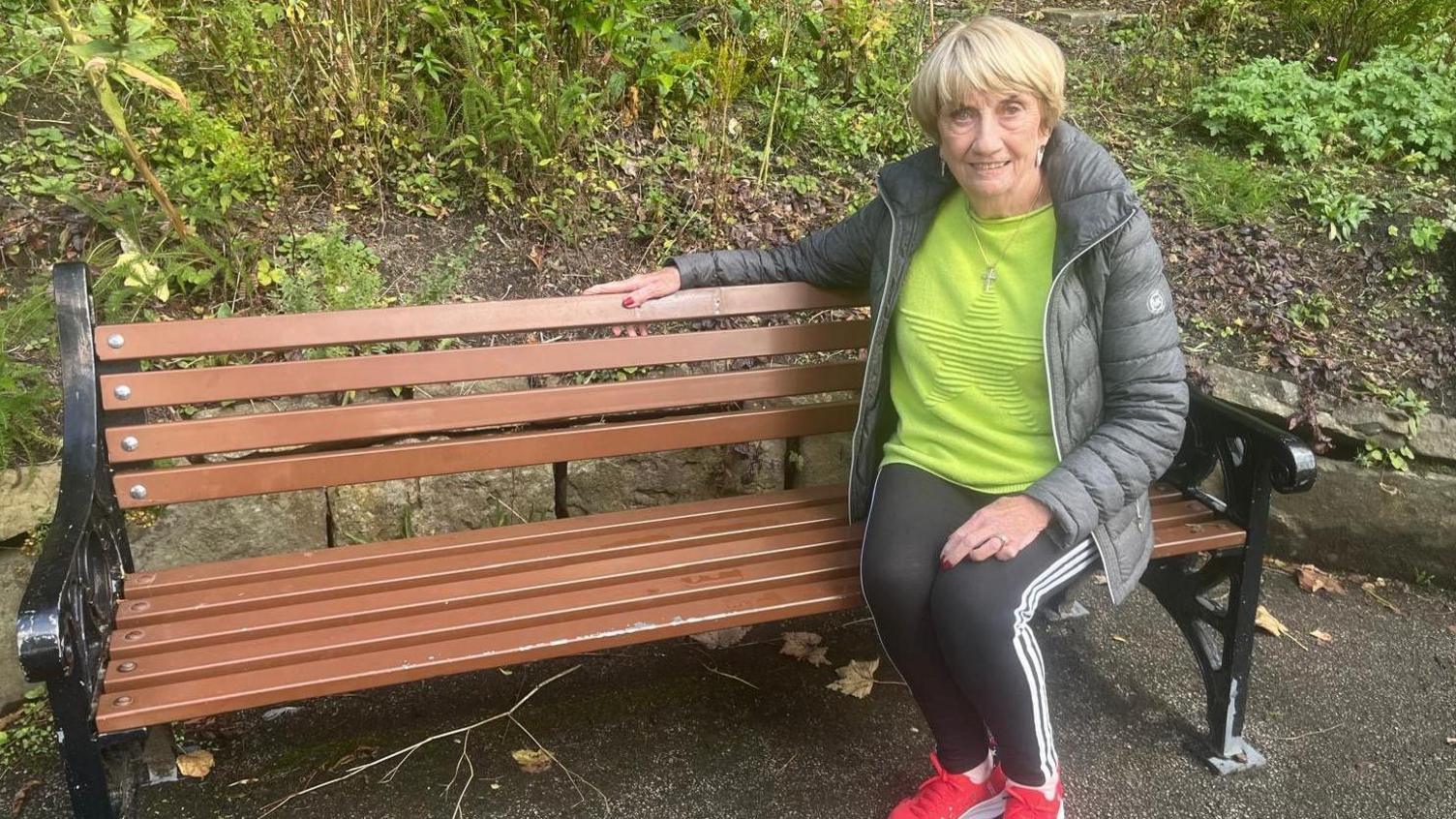 An elderly woman with short blond/grey hair sits on a wooden park bench while smiling. 
