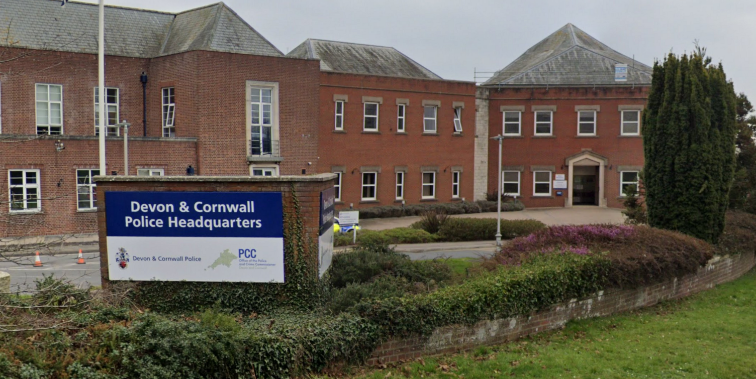 The Devon and Cornwall Police Headquarters building is a series of adjoining red brick, two-storey buildings. There are many windows and the buildings have grey slate roofs. There is a sign at the front which reads "Devon and Cornwall Police Headquarters" and bears the force's logo which is a map of Devon and Cornwall. There is a planted flowerbed in the foreground and a lawn.