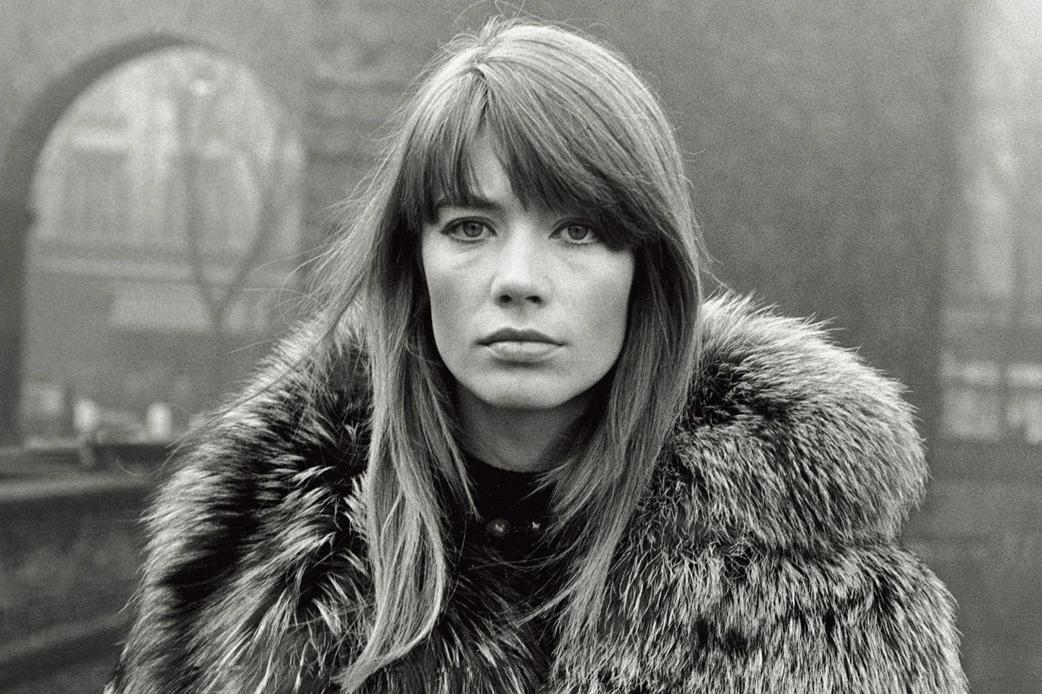 Françoise Hardy photographed in the 1960s on what looks like a winter's day in Paris. She has long brown hair and wears a thick fur coat. She stares directly and seriously at the camera 