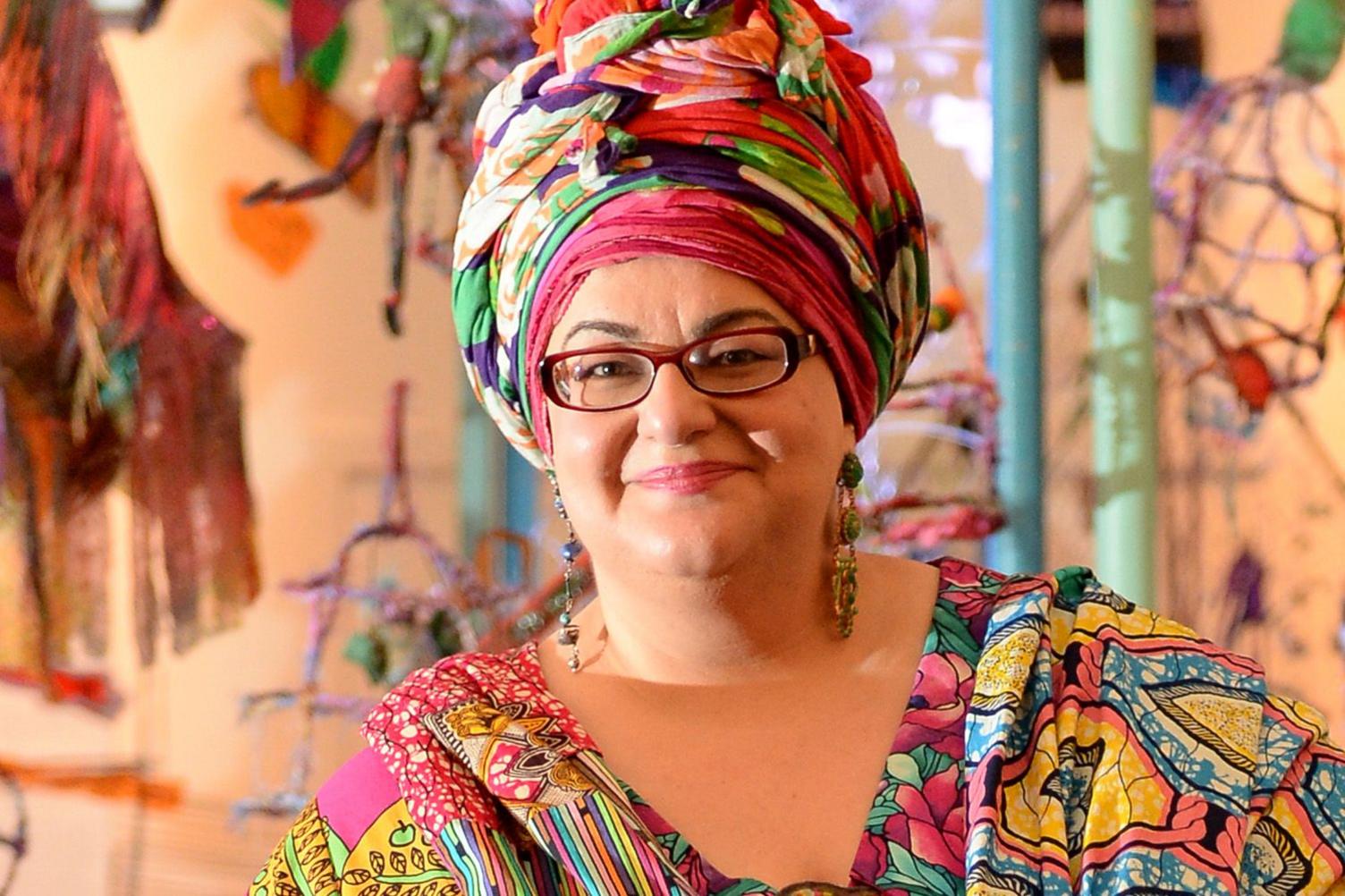 Camila Batmanghelidjh smiles at the camera, wearing glasses. Her clothes are a riot of colour - she has a multi-coloured head scarf and an equally colourful dress and shawl. Behind her appears to be some sort of art installation.