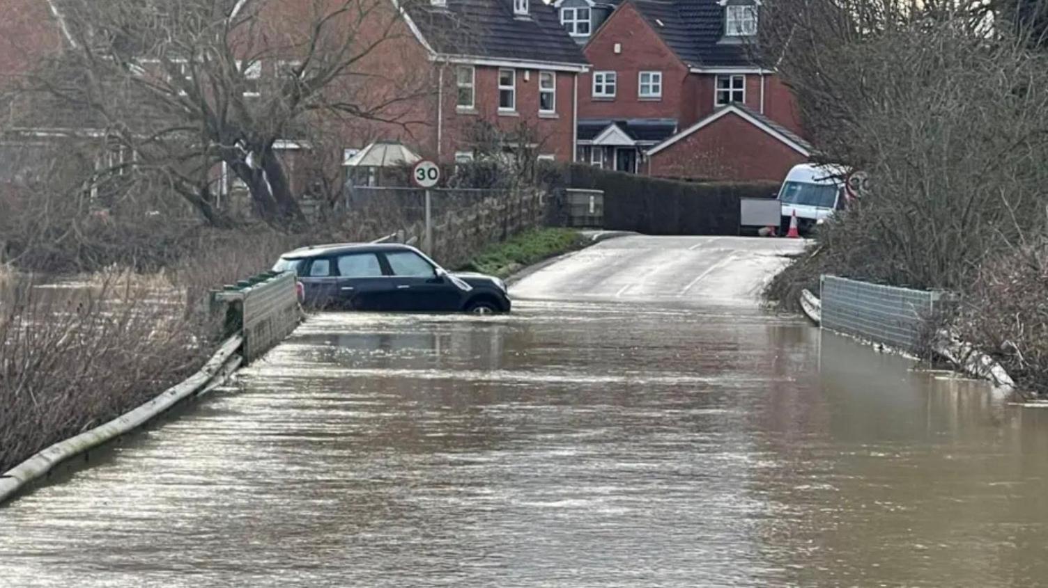 A black mini part-submerged in floodwater on a road over a bridge