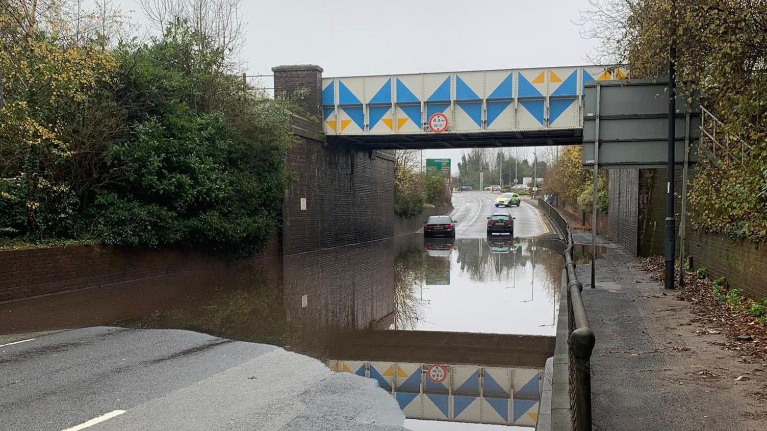 A road under a railway bridge that is flooded with two cars stranded and a police car in the distance.