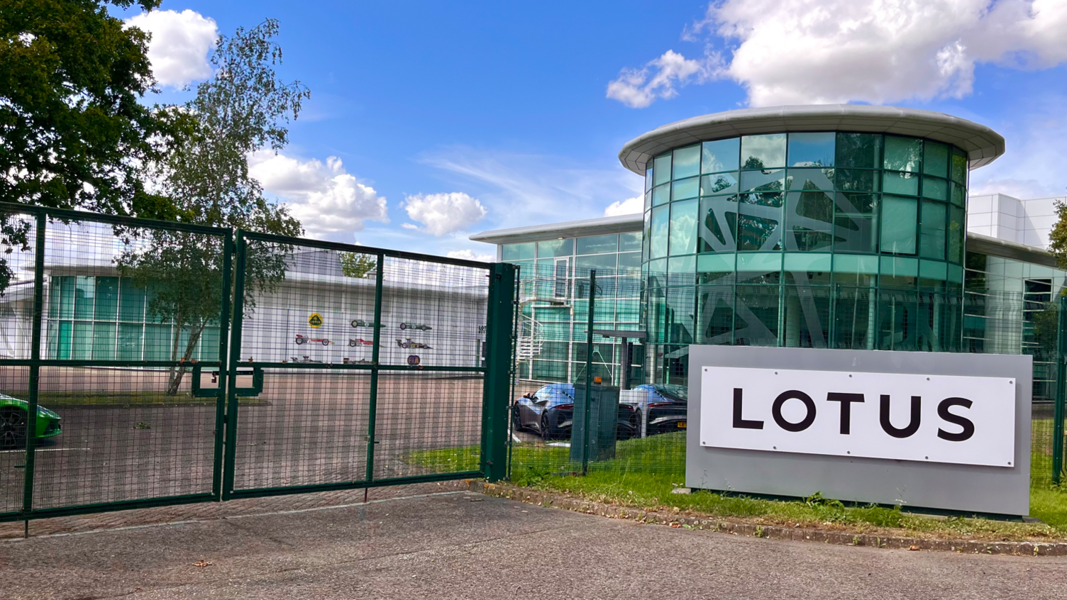 Green metal wire gates in front of a car park, with a glass fronted building to the right and a black-on-white sign that says LOTUS.