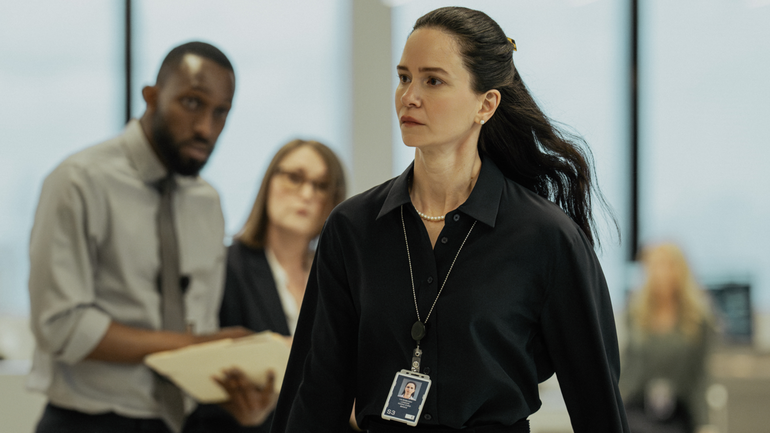 Katherine Waterston walks around an office wearing a black shirt and lanyard with people in the background staring at her