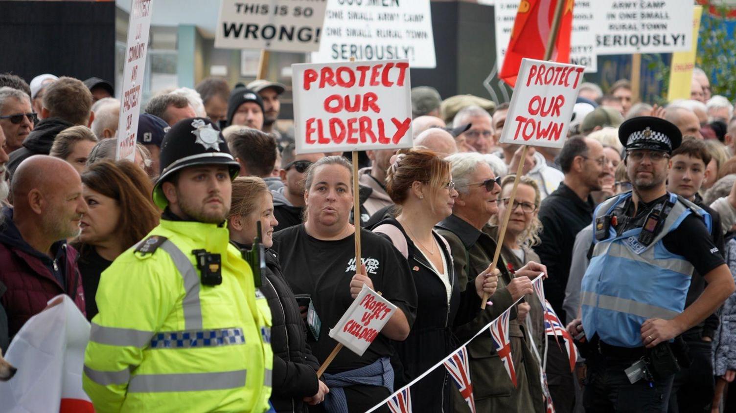 People standing with placards behind a Union Jack bunting. There are two police officers standing next to them.