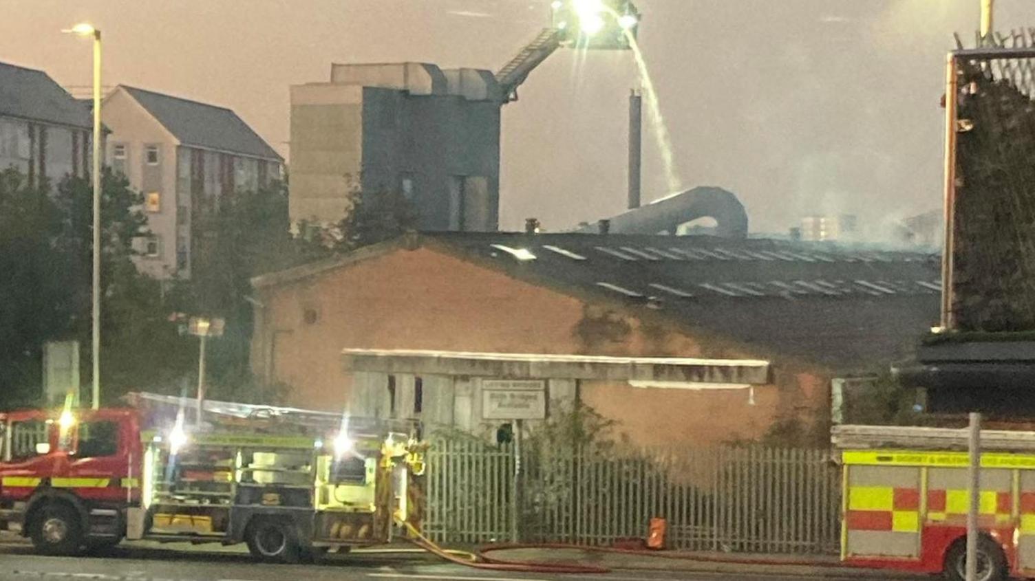 An aerial platform sprays water onto the fire which is obscured by a building. Two fire engines are in the foreground.