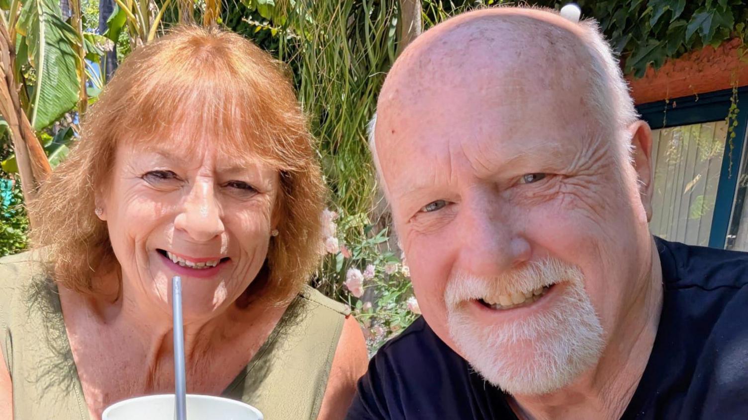 Anita and George Roberts smile at the camera. Anita has shoulder length red hair and holds a cup and straw to her face. George is bald and has a grey goatee.