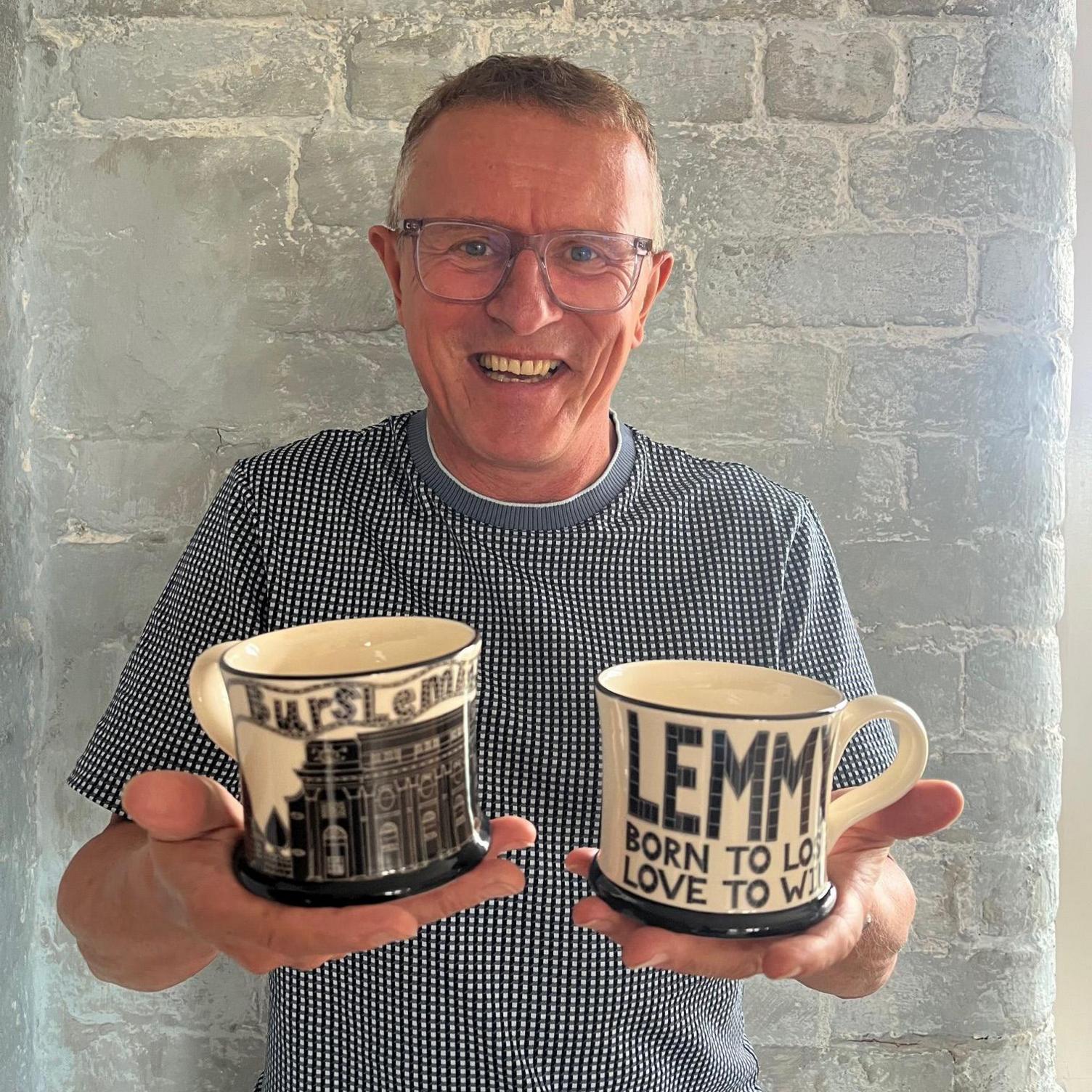 A man with short hair, glasses and a blue and white shirt, smiles as he holds out two of the Lemmy mugs. 