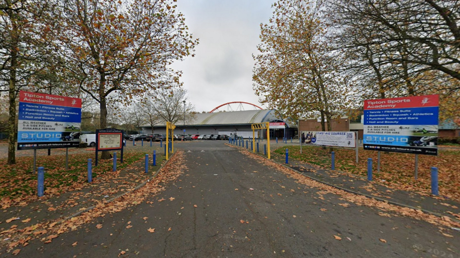 A road leads up to a sports complex in the distance. Signs on either side of the road read "Tipton Sports Academy".