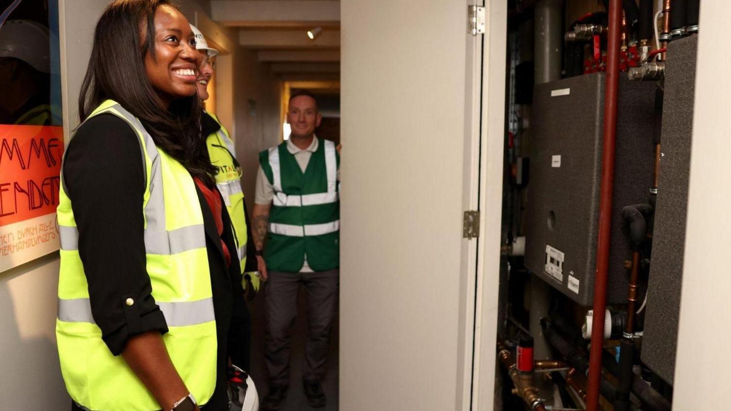 A women in a yellow high viz jacket smiles as she is shown an electricity installation inside a building. There is a man in a green high viz jacket facing the camera.