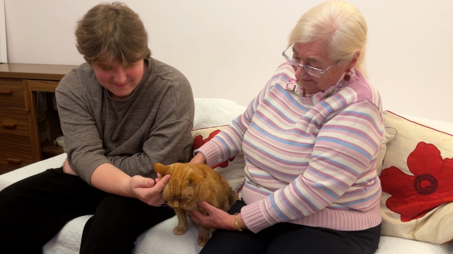 Man with grey jumper on stroking a ginger cat. There's a lady sat to the right on the sofa with a pink, white and blue jumper on.