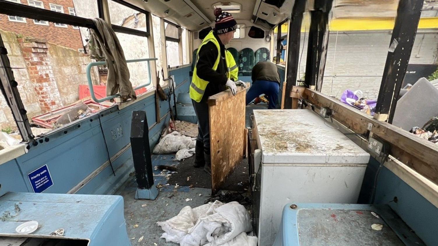 Council workers wearing yellow hi-vis vests clearing debris from the inside of the abandoned bus. The vehicle is blue on the inside and filthy, with torn duvets, rubbish and smashed window glass everywhere. 