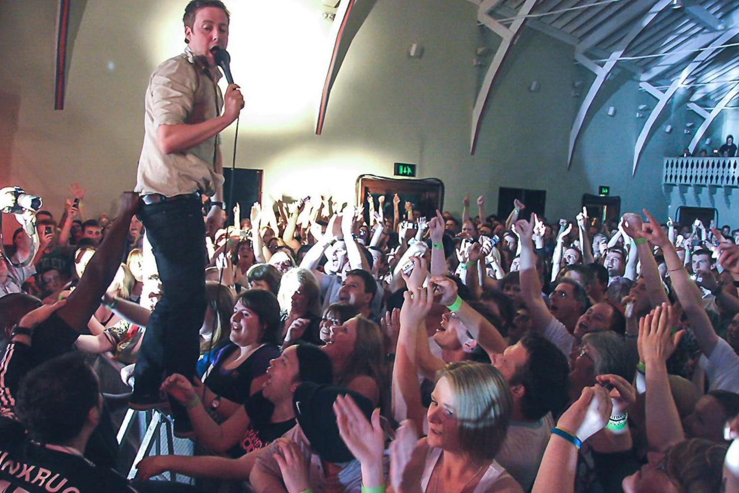 Kaiser Chiefs frontman Ricky Wilson stands on a barrier surrounded by fans.