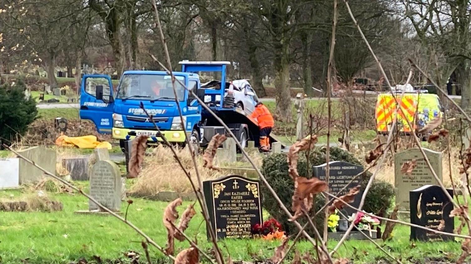 A blue recovery truck with its ramp down and a silver car being winched on to the back of it. There are a number of black and grey gravestones in the foreground.  