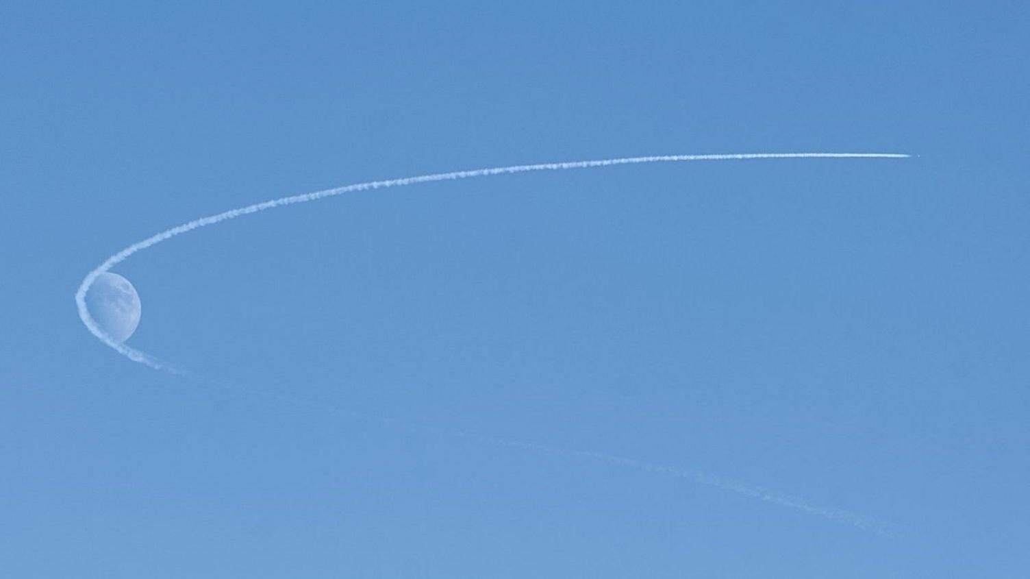 Blue sky and plane trail making an arc around the moon.