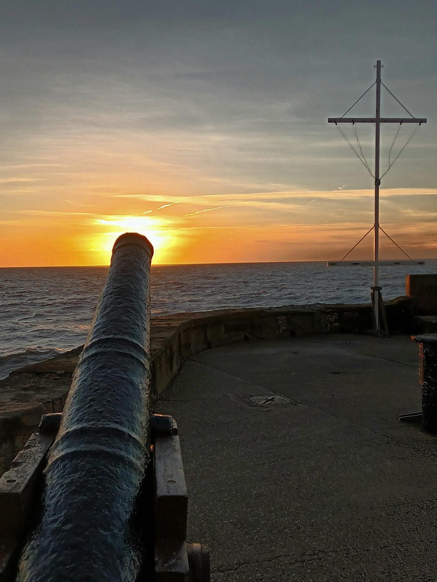 A photograph of a canon overlooking the sea, the placement of the sun is at the end of the canon. The sky is a mix of golden orange and dusty blues. The sea looks dark blue and calm.
