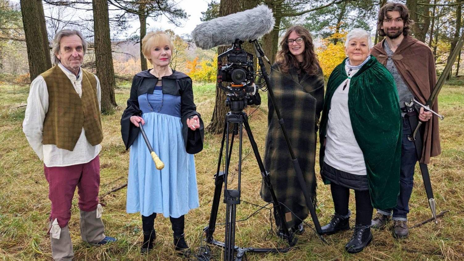Five people stand in a line in a forest. In the centre is a camera and boom mic, with Rosie stood to the right. On either side of her are people dressed as fantasy characters. On the far left, a man wears a large white shirt and a chequered waistcoat and boots; another man on the far right wears a cape and holds a prop sword. Between them, on either side of Grace, stand a woman with short white hair wearing a velvet green cape, and another woman, in a blue dress, holding a wand and wearing a tiara.