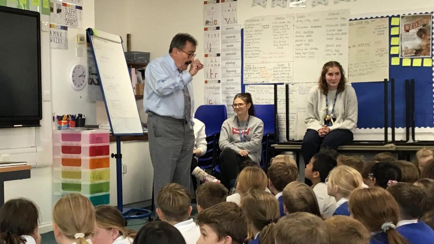 Lord Winston with dark hair and beard, wearing a light blue shirt and grey tie, with one fist raised. Several rows of boys and girls, some with blue school sweaters, are sitting on the floor in front of him. An adult is sitting on a desk, two others are sitting on chairs. There is a whiteboard behind Lord Winston.