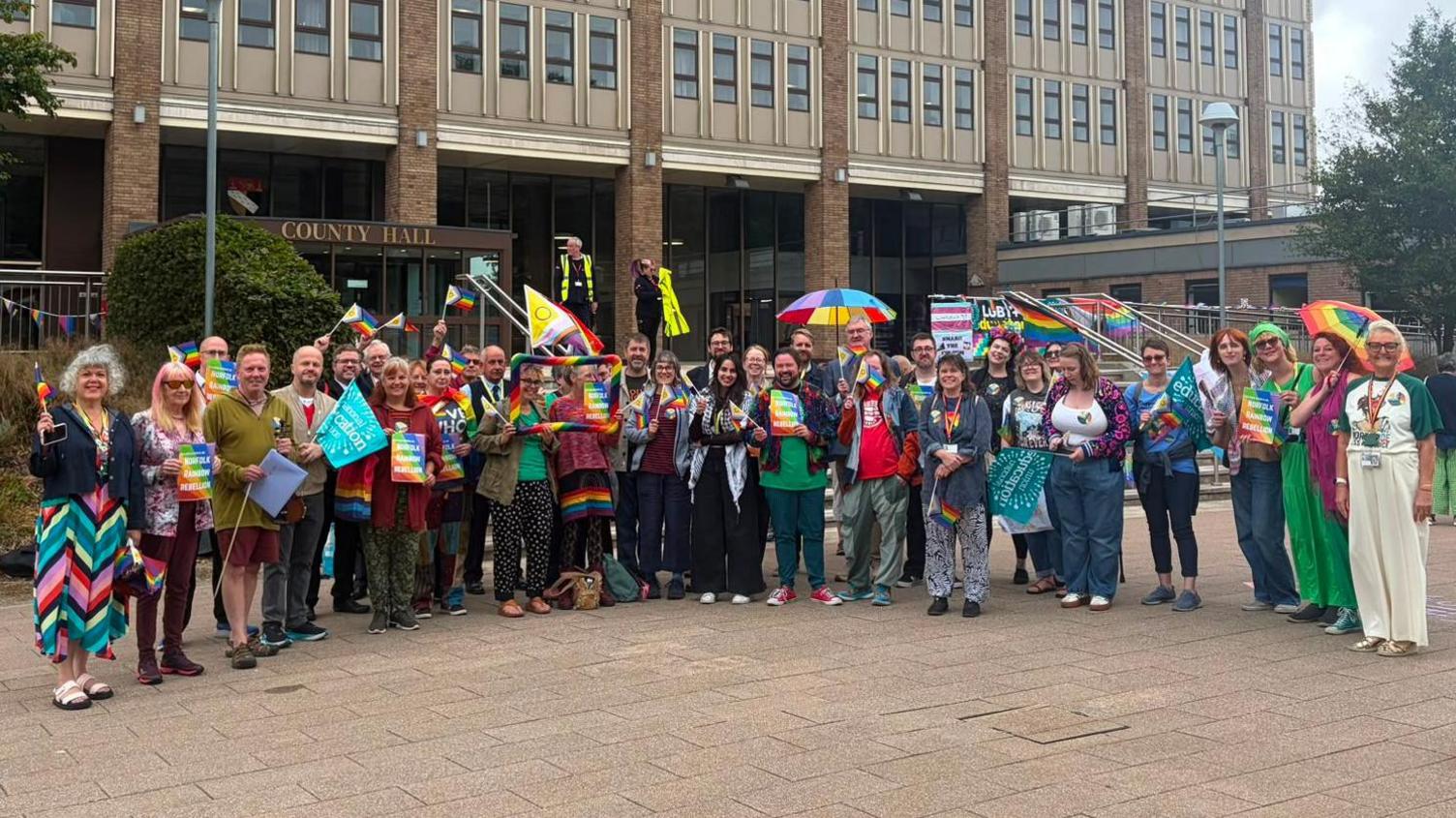 A group of people gather outside County Hall in Norfolk, holding up various rainbow-coloured symbols, flags and placards