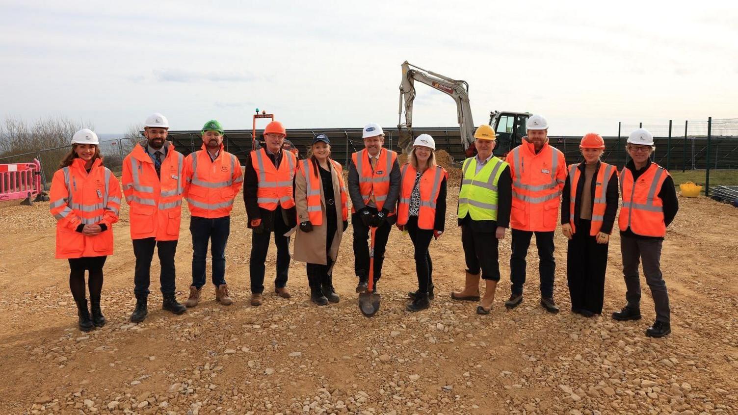 Eleven people standing in a line in hi-vis clothing. A man in the middle is holding a shovel. Behind them is a large digger and the brown earth they are standing on is flat and covered in small partially buried rocks.