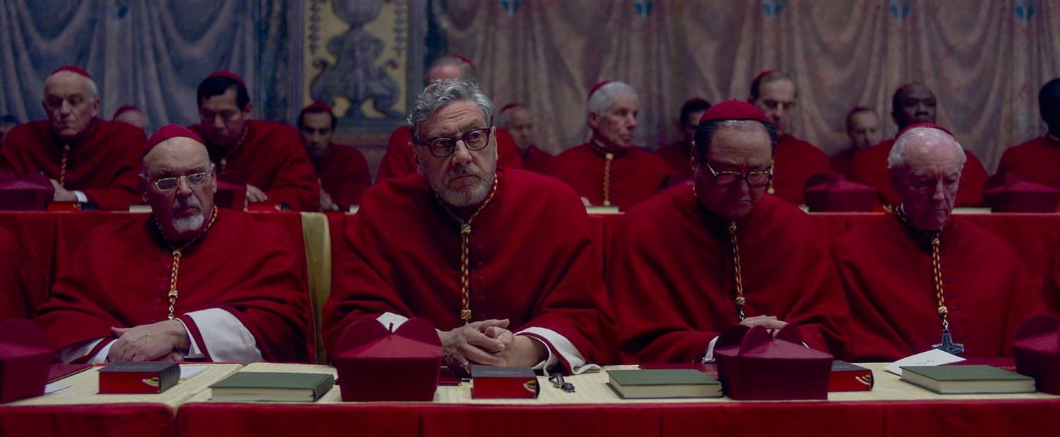 A row of cardinals wearing crimson robes, sat at a table