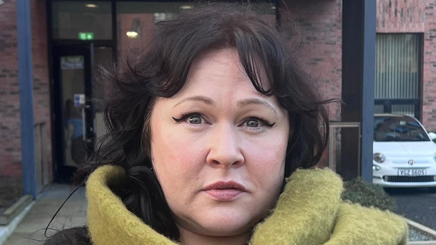 Sara Morrison is wearing a brown top with a large green scarf around her neck. She is standing in front of a red brick building with black windows. There are cars and a motorbike in a car park between her and the building. The path around the building is of white stones. 