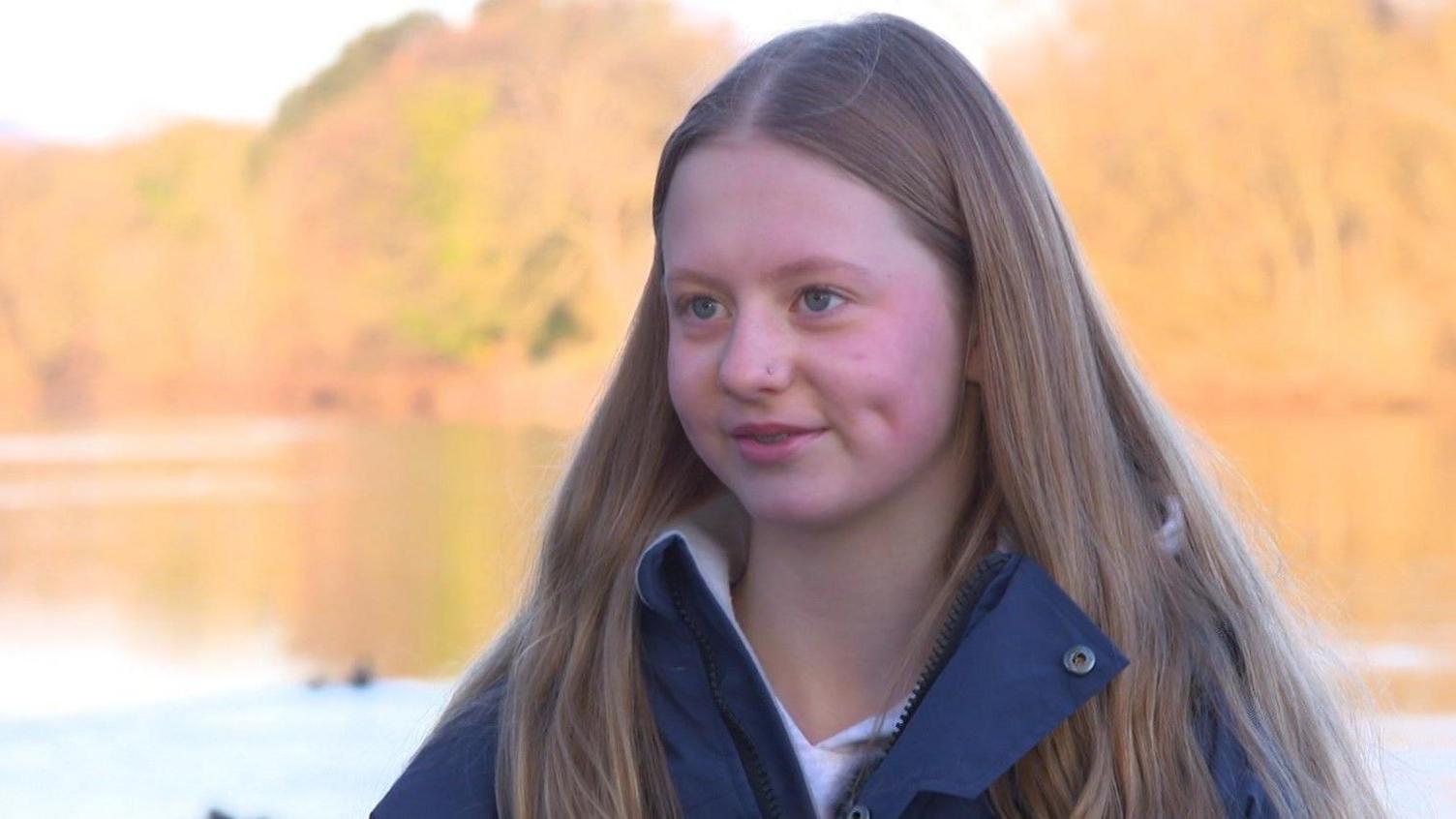 A headshot of Frankie, with the lake in the background. She has long blonde hair, which is wavy at the ends and parted in the middle. Her eyes are blue and she has dimples. Her cheeks are rosy from the cold. 