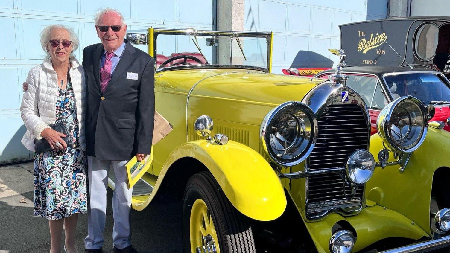 Malcolm Stern and his wife pose next to the completed car, which is bright yellow with large head lamps. He is wearing a suit and sunglasses. Both are smiling.