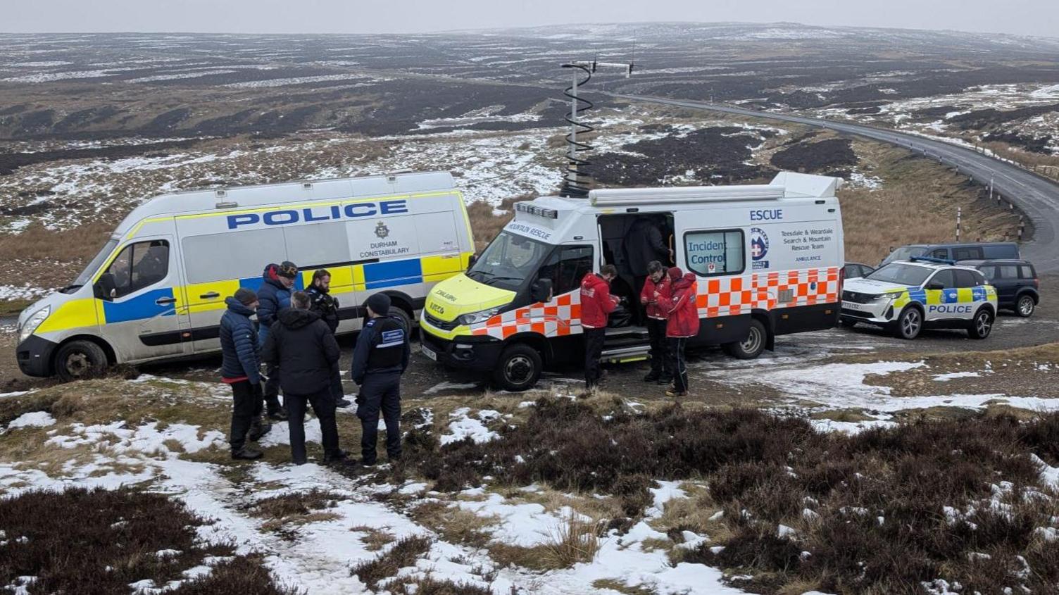 A police van parked alongside a mountain rescue van. Mountain rescuers can be seen in front of the vans in red jackets with police officers in black standing in a separate group. Moorland can be seen behind the vans, which stretches beyond the horizon.