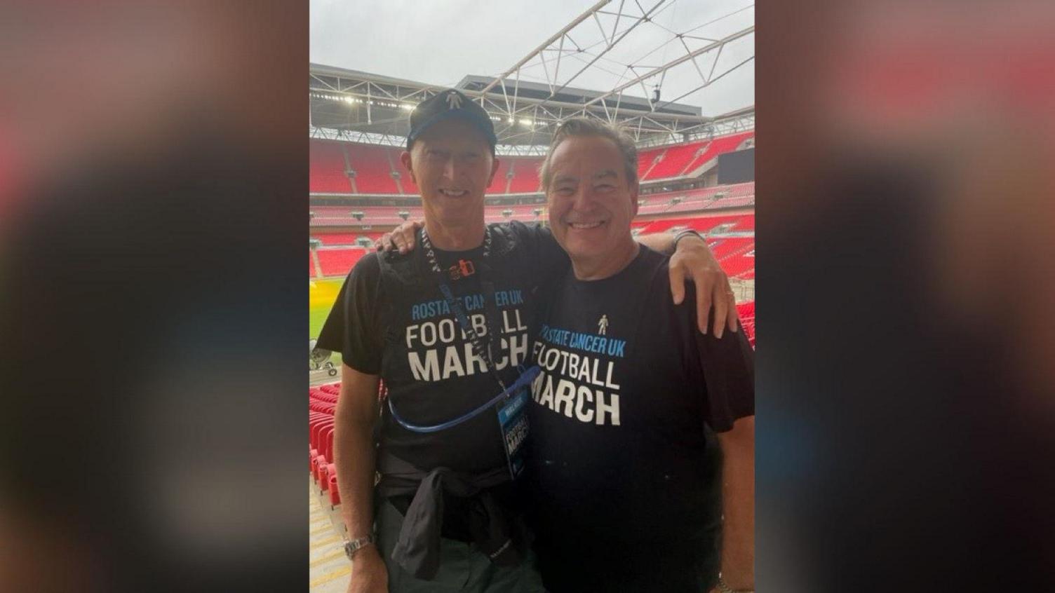 Paul Dennington and Jeff Stelling look into the camera and smile while embracing each other. They have arms round each other and are both wearing tops for Prostate Cancer UK.