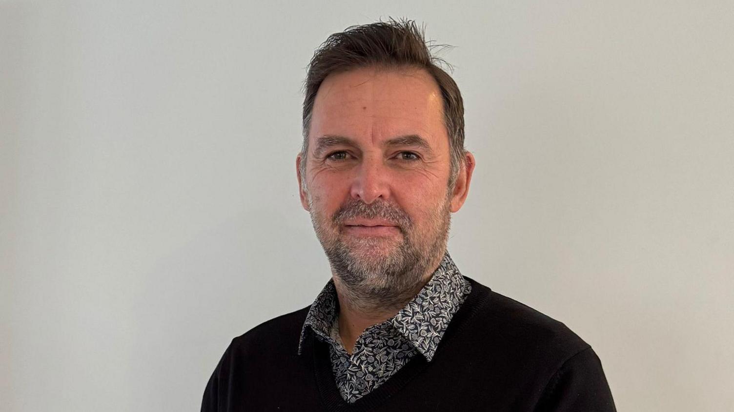 Andre Austin is a man with dark hair and a grey beard. He is wearing a black jumper and a black and white shirt. The background is a white wall.