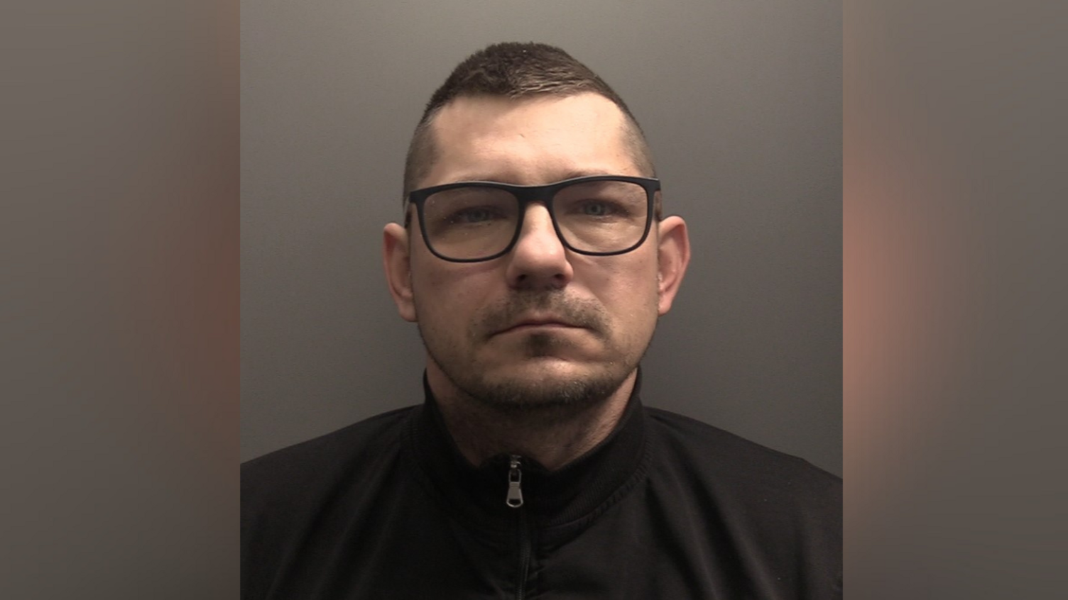 Man with short brown hair and glasses with dark frames. He is wearing a black top with a zip.