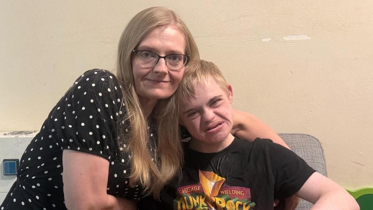 Michelle, a woman with blonde hair, is wearing a black and white dotted dress, kneeling next to her son. He is wearing a black T-shirt and looking at the camera.