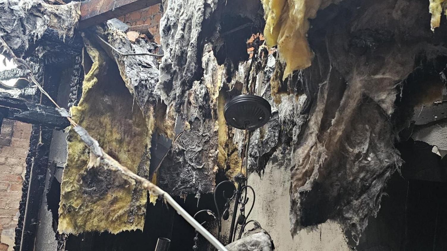 Image shows the remains of a wall in a property after a fire. Blackened items badly damaged by fire along with yellow insulation hanging from the walls and roof of the building