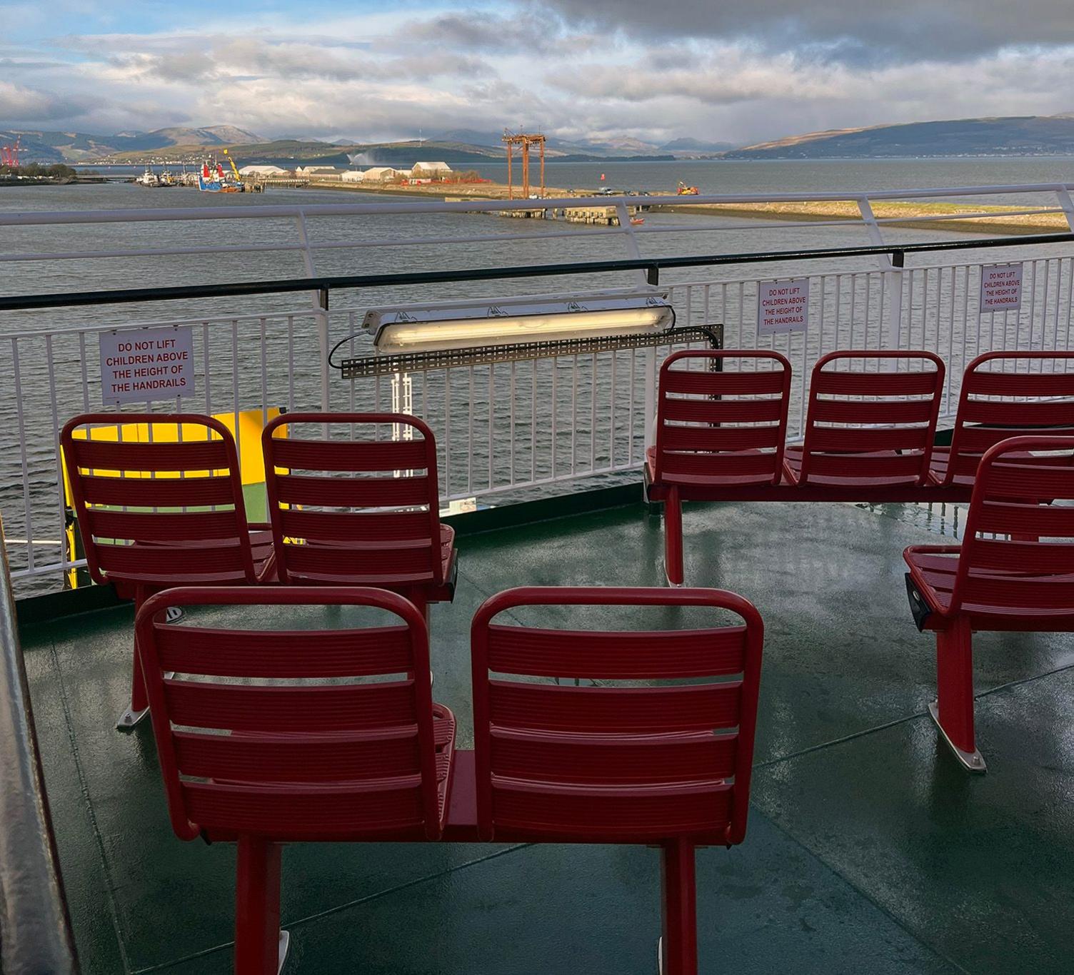 Red metal seats with slatted backs, facing out over a safety railing in the external seating area of the ship