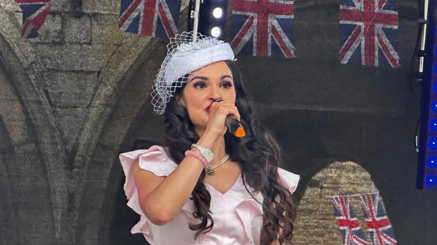 A performer singing on a stage at armed forces day. There is Union Jack buntain behind her/ She is wearing a light pink dress, fascinator on her head and has long brown hair. She is holding a microphone in her hand.