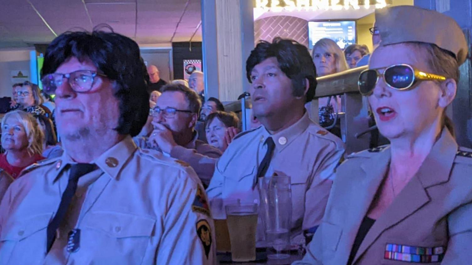Two men and a woman dressed in beige US military uniform like the ones Elvis worn during his military service. The men both have elvis wigs on while the woman is wearing dark glasses with gold rims like Elvis. 