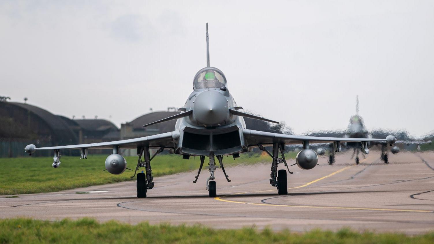 The front of a taxiing Typhoon jet on the runway at RAF Coningsby in Lincolnshire with a second plane and hangars and a grassy area in the background. 