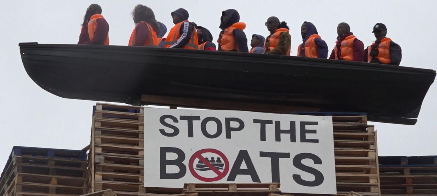 The picture shows a model of a small boat with figures inside in orange fluorescent vests. Below is a sign which says 'STOP THE BOATS'