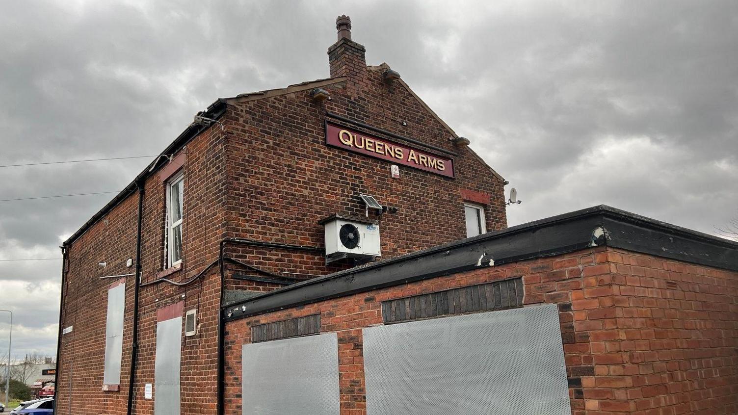 A former pub with boarded-up windows. A red-and-gold sign on the side of the brick building reads "Queens Arms".