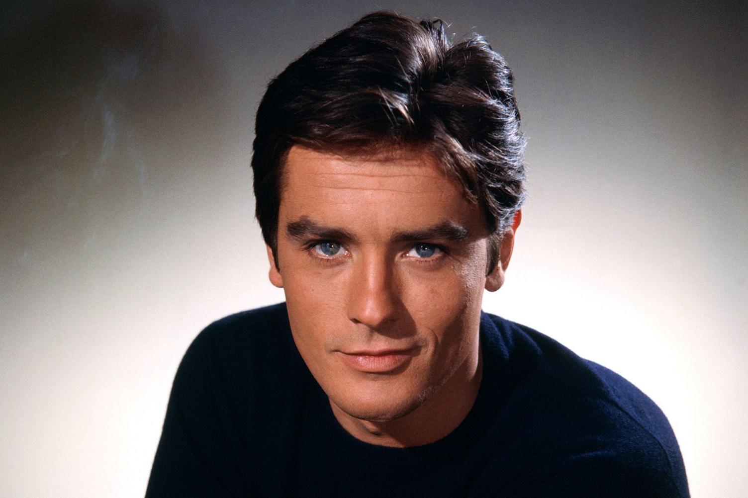 Undated 1960s publicity photo of Alain Delon (in colour) when he would have been in his late 20s or early 30s. He has dark, slightly bouffant hair,  clear blue eyes and is smiling slightly. He wears a black sweater and is looking straight at the camera. 