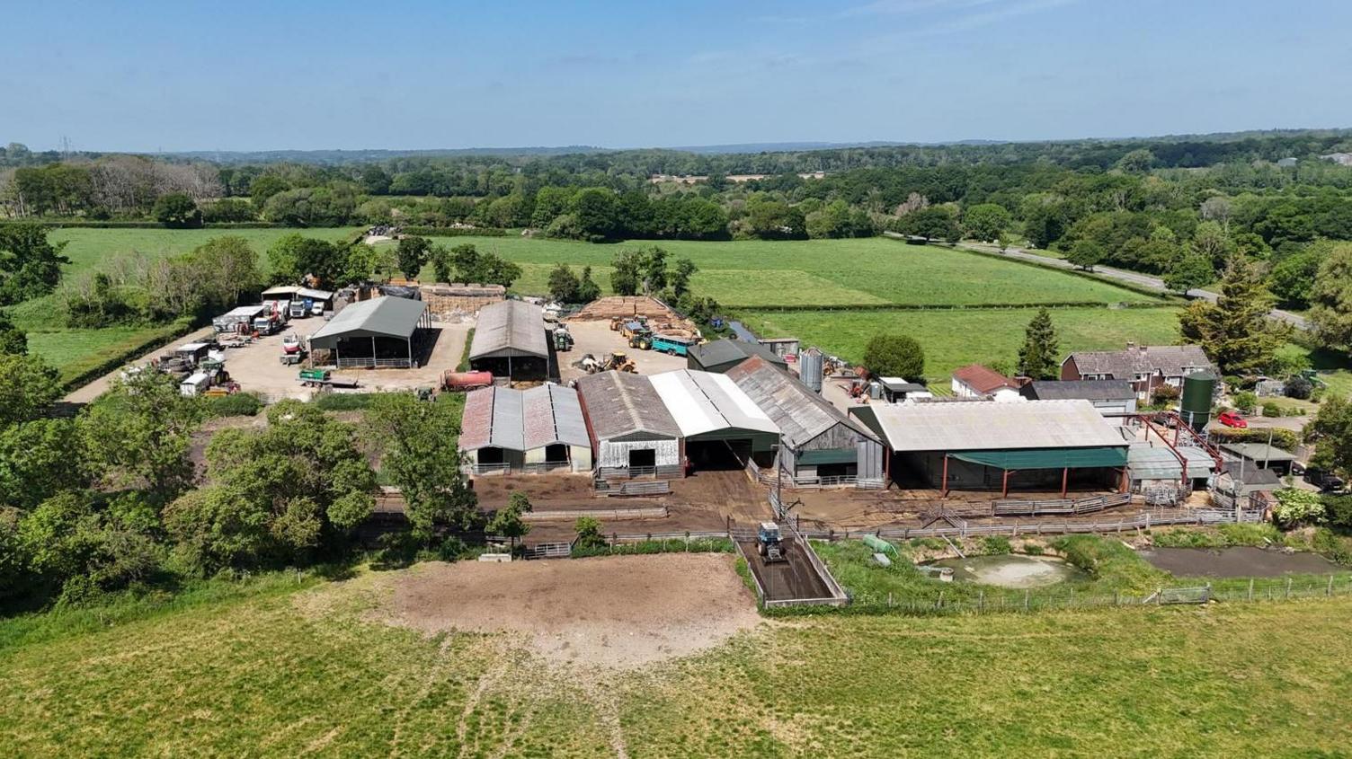 Aerial view over Thistle Ridge Farm - a collection of barns and machinery surrounded by field and countryside with a road running along the right side of the picture. There are three residential buildings to the right of the farm near the road.