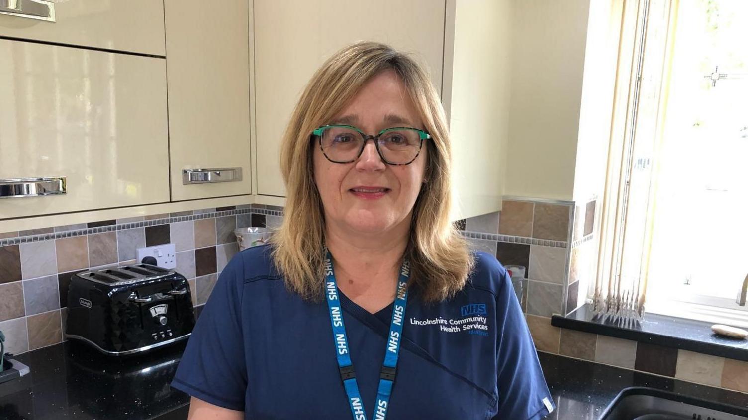 Julie Jarvis is looking directly at the camera and smiling. She is standing in her kitchen. She is wearing navy scrubs and a light blue NHS lanyard. She has green rimmed glasses. "NHS Lincolnshire Community Health Services" is embroidered on her top.